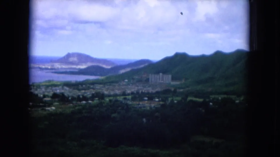 1964 Hawaii 8mm frame with a coastal town below mountains, blue water beyond, and a Diamond Head-like form in the distance.