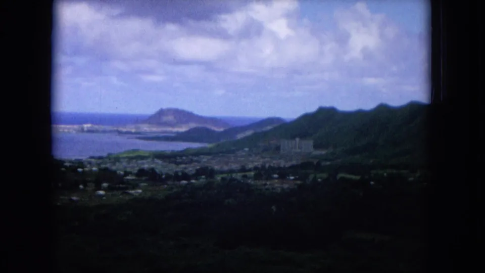 1964 Hawaii home-movie view across foreground vegetation to a coastal settlement, water, mountains, and distant ocean horizon.