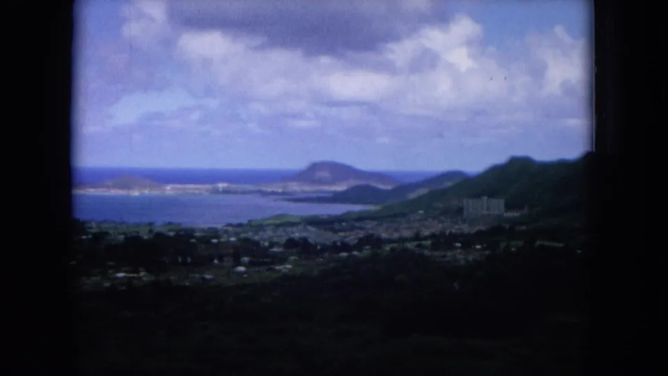 1964 Hawaii 8mm frame looking over a coastal town and blue water toward a rounded Diamond Head-like landform under clouds.