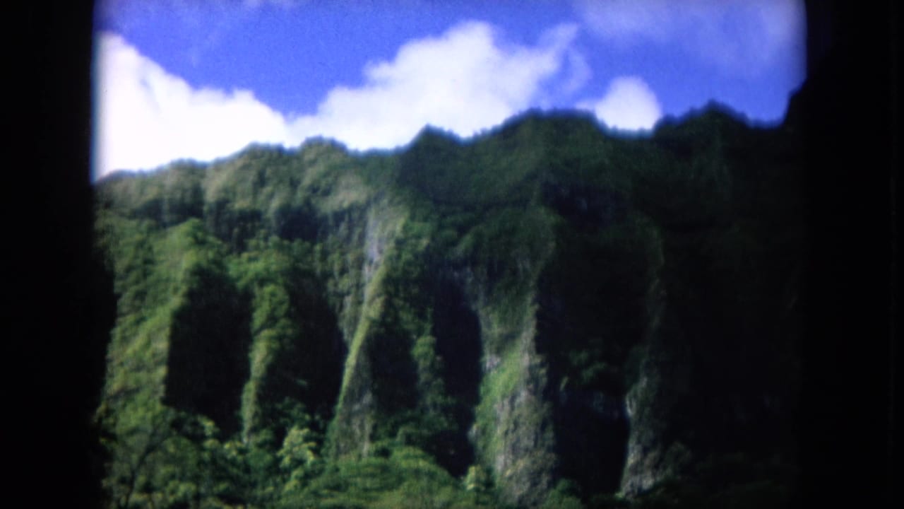 1964 8mm home-movie frame of a lush Oahu mountain ridge, dense greenery, and bright clouds seen through dark film edges.