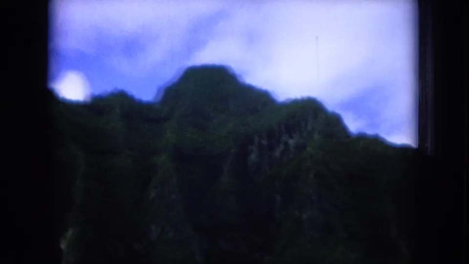 Soft 8mm frame from 1964 Oahu footage, showing a shadowed green mountain slope beneath a pale blue sky within dark frame borders.