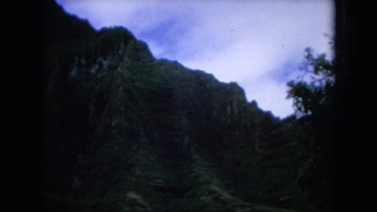1964 Hawaii home-movie frame showing a shadowed green mountain ridge and dense tropical foliage beneath a bright sky.