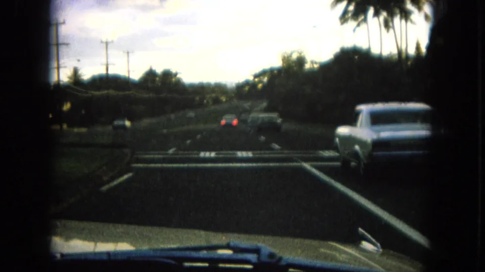 1964 Hawaii home-movie driving view from inside a vehicle, with cars ahead on a multi-lane road lined by palms and utility poles.