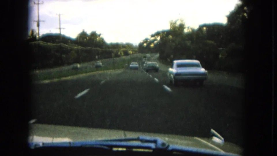 1964 Hawaii highway driving shot from a car, showing several vehicles ahead on a divided road bordered by tropical greenery.