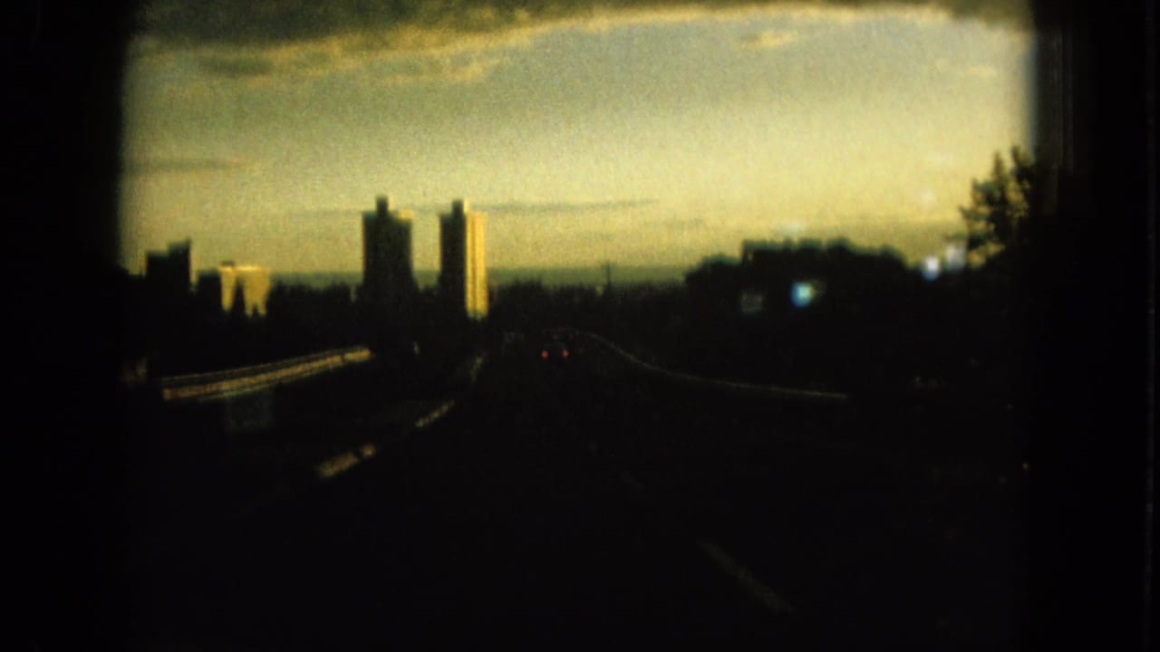 1964 Oahu home-movie frame from a moving car, showing a dark road, guardrails, trees, and distant high-rise towers in warm evening light.