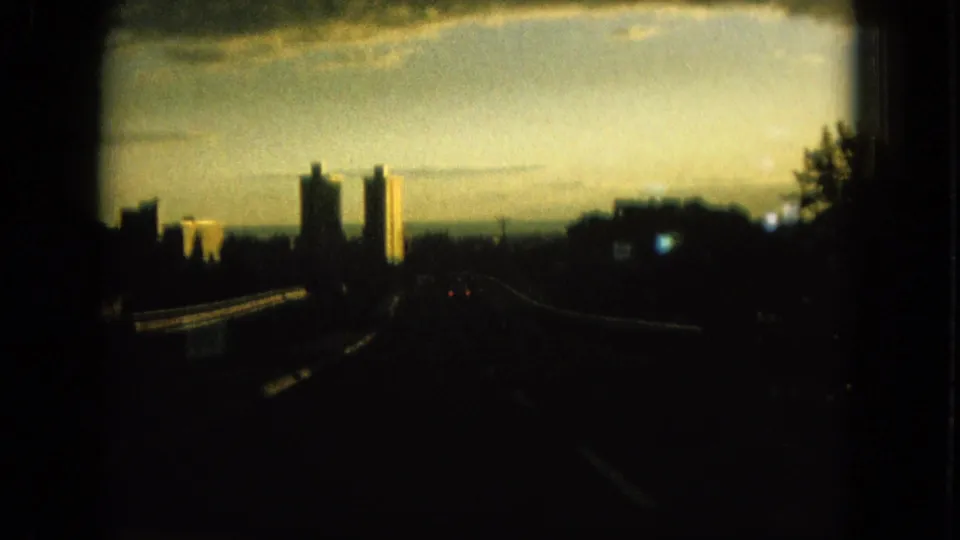 1964 Oahu home-movie frame from a moving car, showing a dark road, guardrails, trees, and distant high-rise towers in warm evening light.