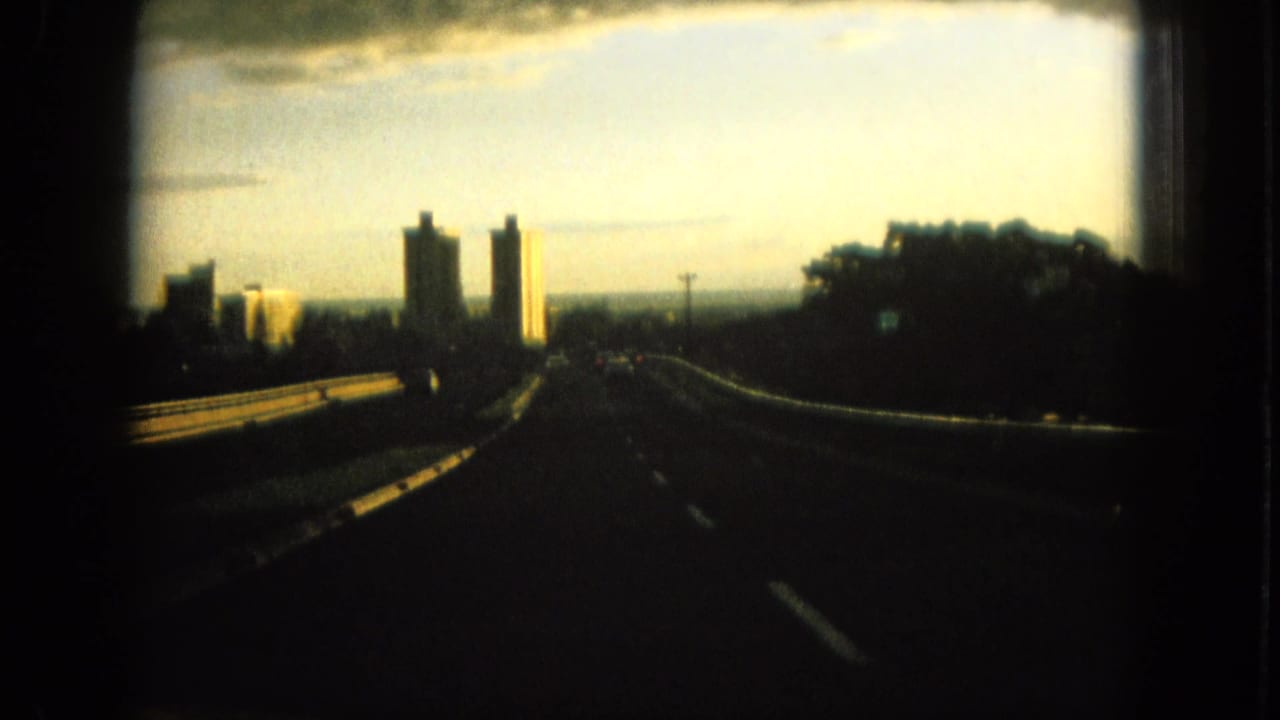 1964 Oahu travel footage frame with a road curving toward hotel-like towers, roadside trees, guardrails, and a pale sunset horizon.