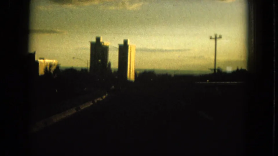 1964 Oahu moving-car frame showing two tall high-rises ahead, a roadside utility pole, guardrail, and silhouetted trees under warm sky.
