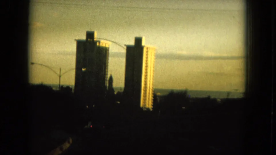 1964 Oahu home-movie frame from the roadway, showing tall high-rise buildings, streetlights, dark foreground, and a golden evening sky.