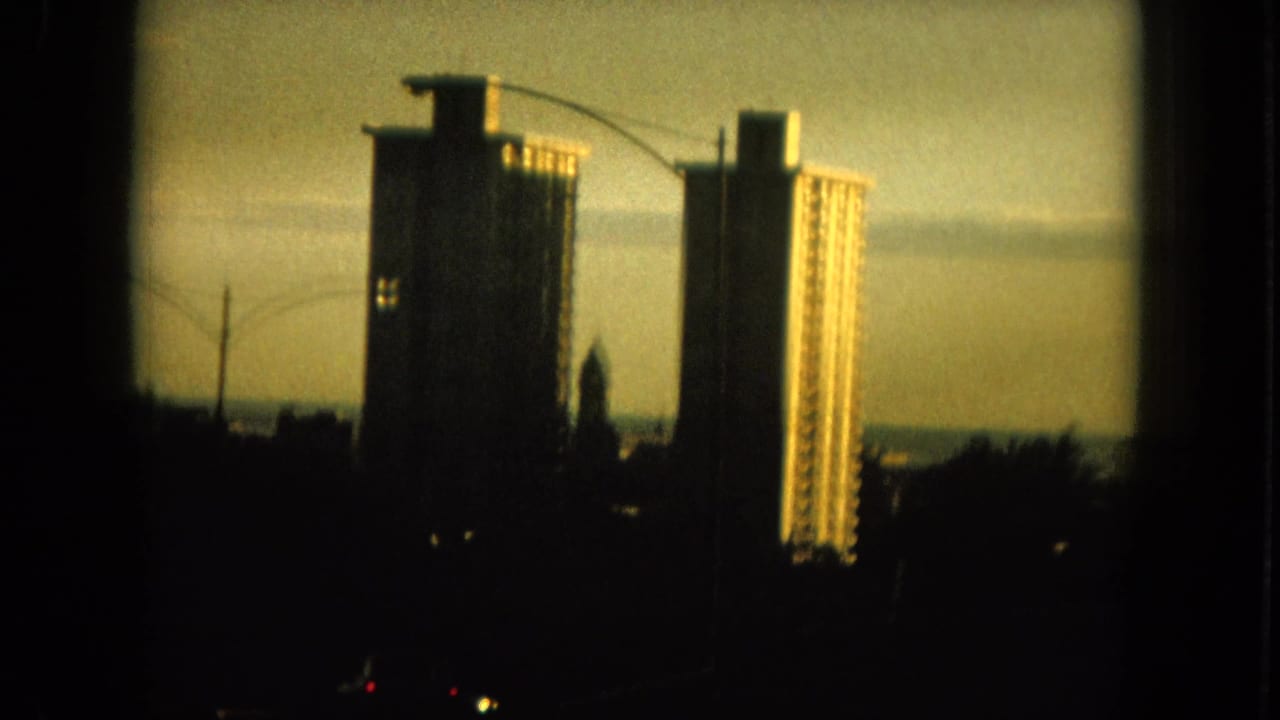 1964 Hawaii home-movie frame showing tall urban buildings in shadow against a hazy warm sky.
