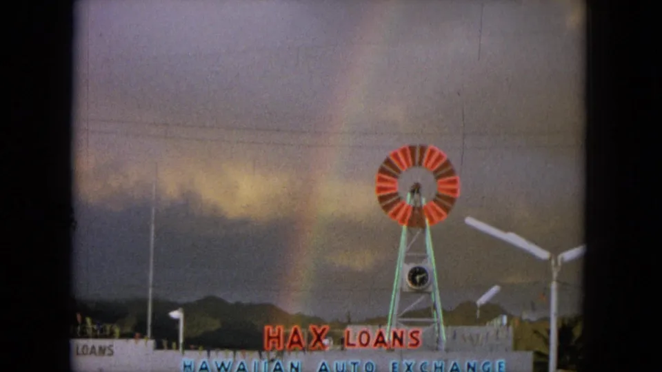 1964 Oahi, Hawaii frame showing a red-and-white windmill, commercial signage, hills, clouds, and a faint rainbow.