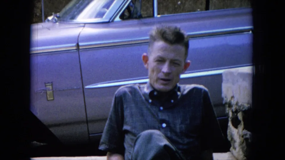 A man in a short-sleeve shirt sits near a large mid-century car, with stonework visible at the edge of the frame.