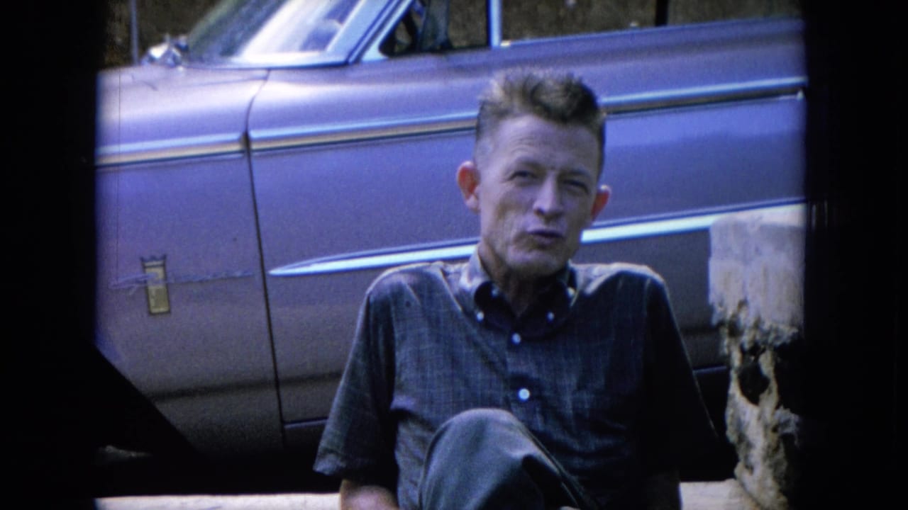 Man seated outdoors beside a vintage car with concrete blocks nearby.