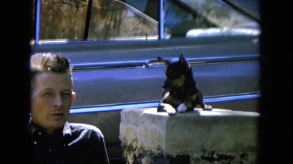 A small black-and-tan dog sits alert on a concrete block, with the man's face partly framed near a vintage car.