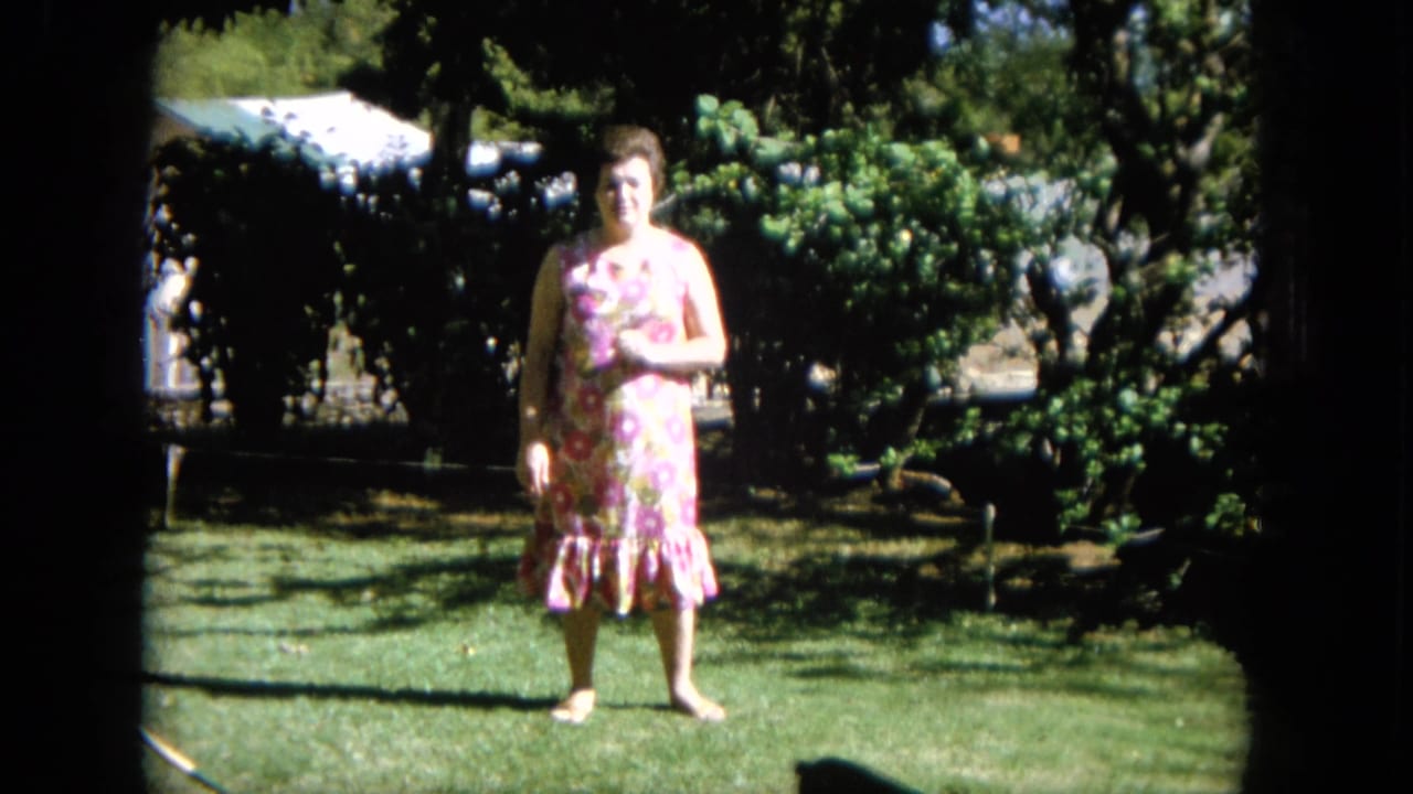 Woman in a floral dress stands centered on the grass with shrubs and tree shade behind her.