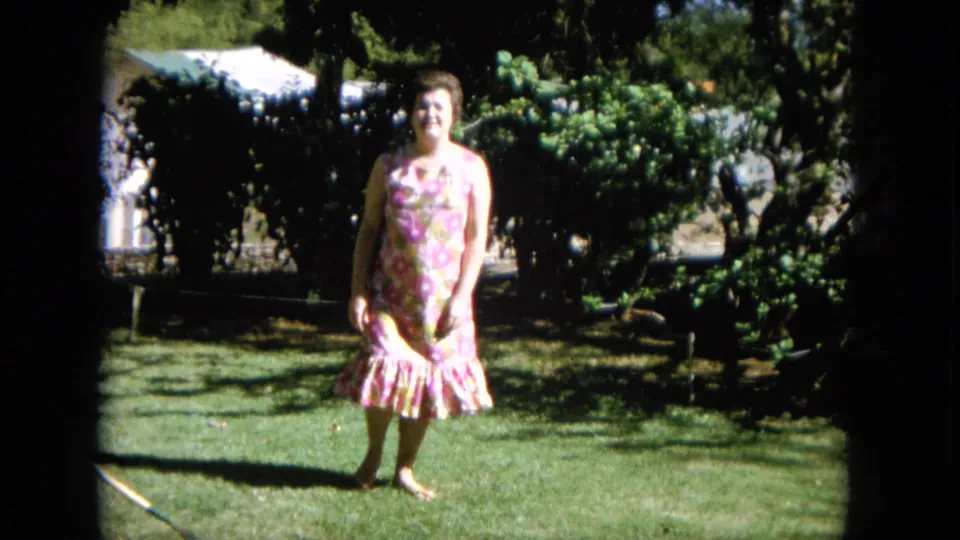 The woman approaches the camera across the lawn, with the floral dress and garden foliage visible in the 1964 Hawaii footage.