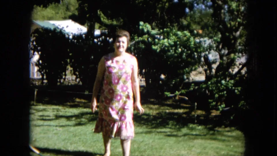 A smiling woman in a pink floral dress walks through a leafy yard in supported 1964 Hawaii home-movie footage.
