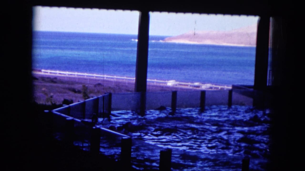 Fenced tidal pool in shadow with blue ocean and shoreline beyond.