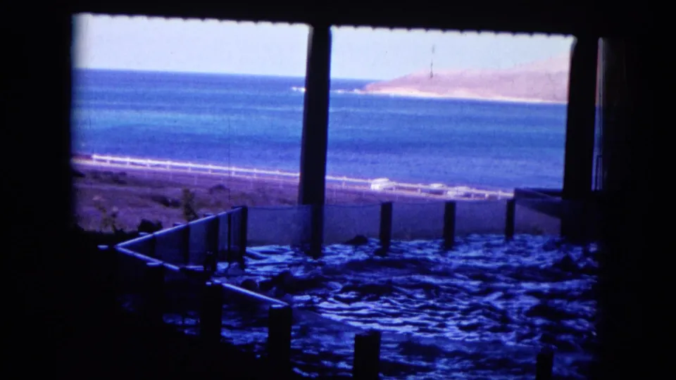 1964 Oahi, Hawaii 8mm frame showing a fenced tidal pool in shadow below a bright ocean view.