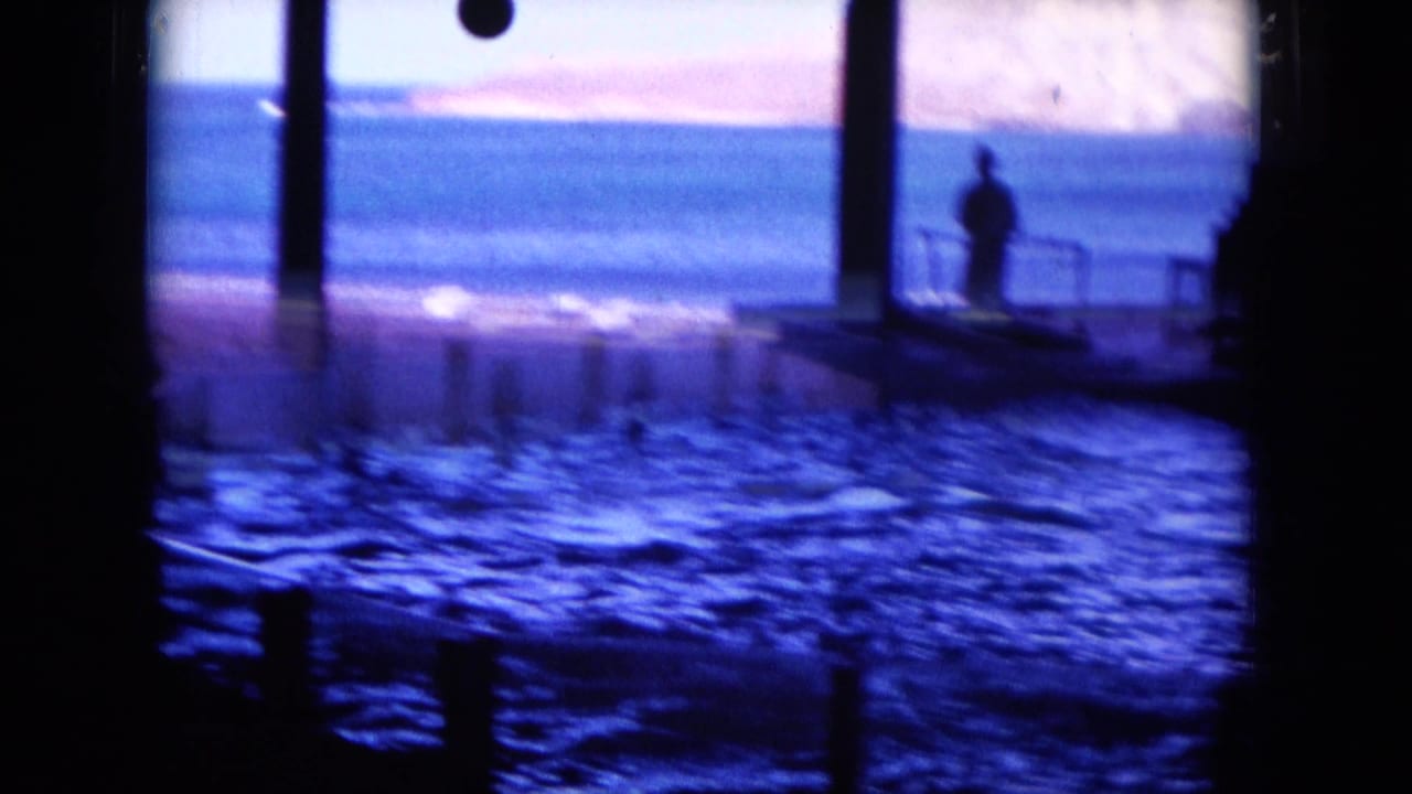 Silhouetted figure stands on a platform beside rippling pool water.