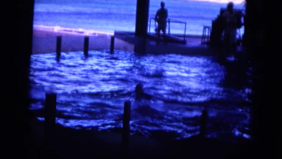 1964 Oahi, Hawaii 8mm frame showing a swimmer in the fenced tidal pool and silhouetted figures by the ocean platform.