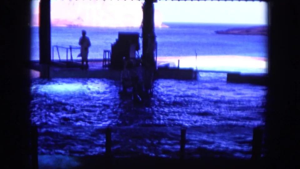 1964 Hawaii 8mm frame showing a person standing on a rail-lined platform beside a blue marine enclosure, viewed through shadowed framing.