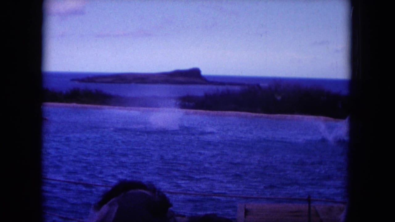 Blue ocean and distant island silhouette seen past a boat rail, with a person partly visible in the foreground.