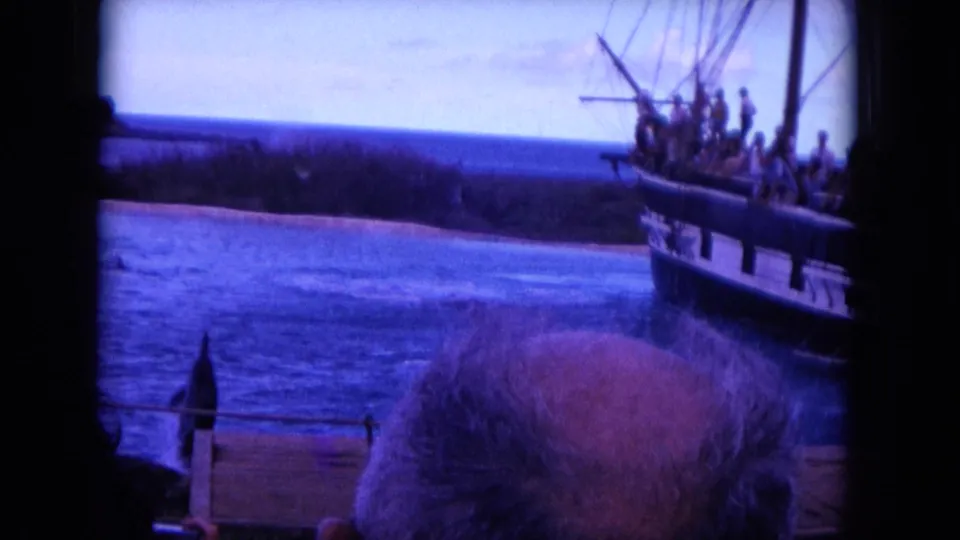 1964 Oahu home-movie frame showing a sailing vessel with passengers near shore, seen past a foreground adult on a boat.