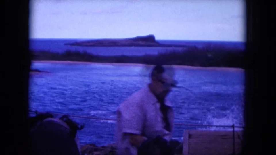 An adult man stands or walks on a boat in 1964 Oahu footage, with blue water and a distant island silhouette in the background.