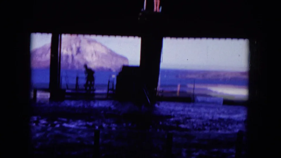1964 Hawaii 8mm frame showing a silhouetted figure beside waterfront railings, with the ocean and distant land visible beyond shaded posts.