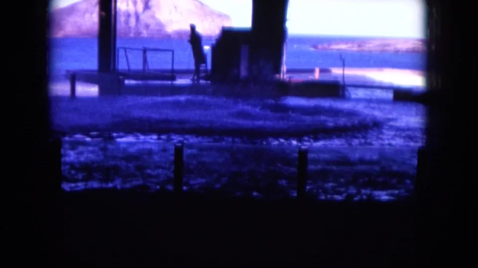 1964 Hawaii 8mm frame of a silhouetted person near a waterfront platform, with blue ocean and a distant island or headland beyond.
