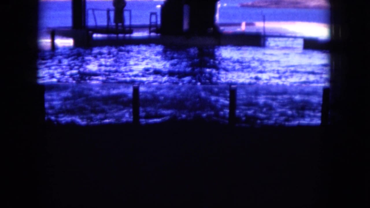 Dark dock structures frame rippling blue water with a silhouetted figure in the distance.