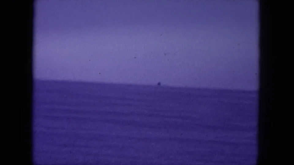 1964 8mm home-movie frame showing calm ocean, distant horizon, and visible gate edge in a purple-shifted scan.