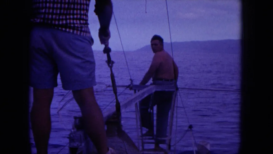 1964 Oahi, Hawaii 8mm frame with two adult men aboard a small boat, one shirtless and one in shorts near rigging.