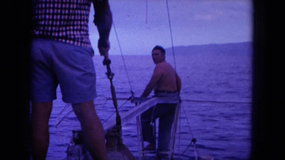 1964 Oahu home-movie frame showing two adult men on a small boat, one seated or crouched near the bow and another standing in shorts.