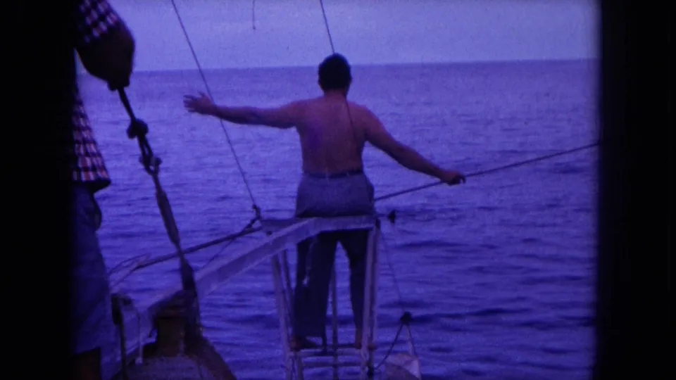 1964 Oahu frame of a shirtless man balancing on a small boat bow with one arm out and a long pole extended over the ocean.