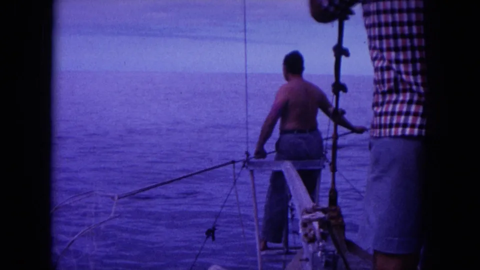 1964 Hawaii home-movie frame with a shirtless man looking over the stern as fishing gear and calm ocean fill the view.