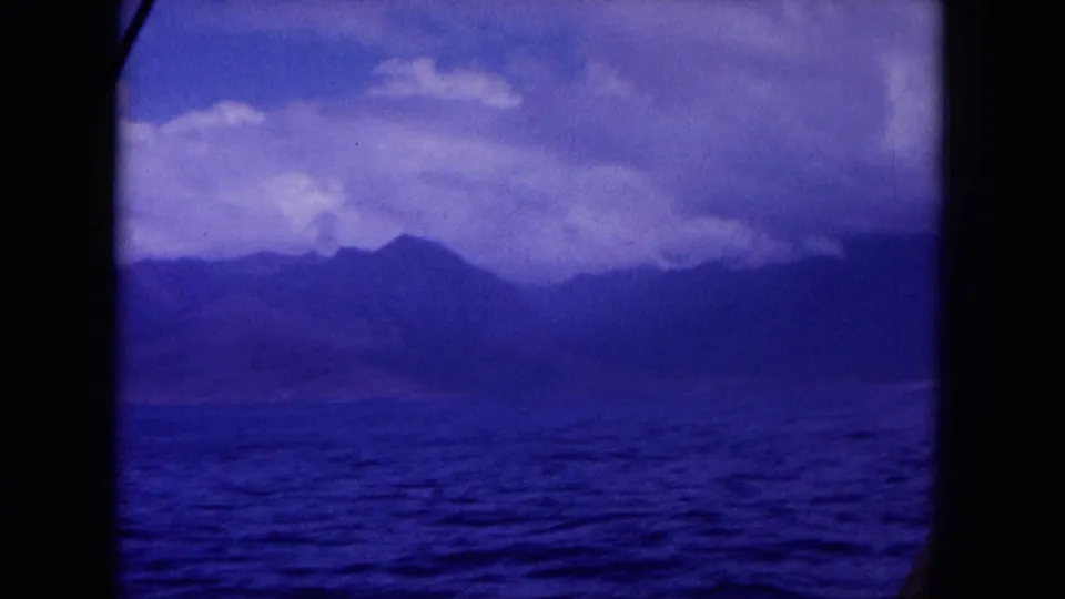 1964 Hawaii home-movie view of a mountainous coastline across the water, seen through a dark boat window frame.