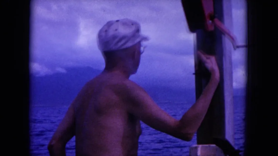1964 Hawaii home-movie frame of a shirtless adult in a white cap beside a boat opening, with water and mountains beyond.