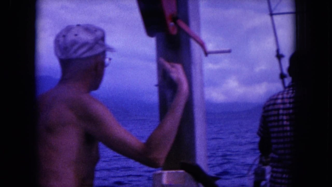 Shirtless man in cap stands on a boat beside another man facing the ocean.