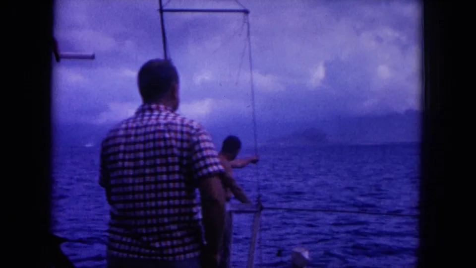 1964 Hawaii home-movie frame showing a man on deck while another person gestures toward the ocean and distant land.