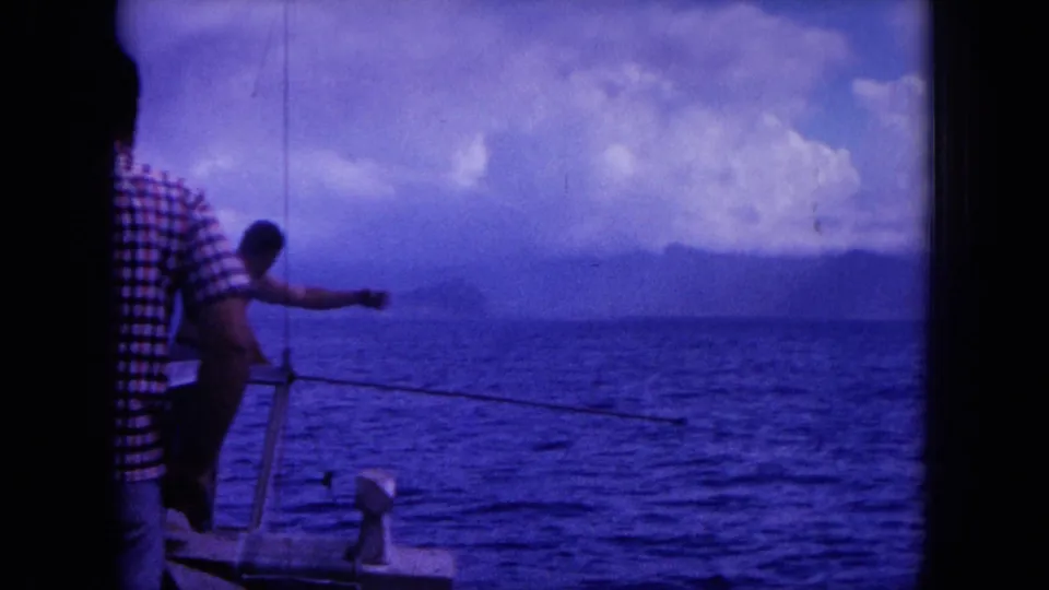 1964 Hawaii 8mm frame of men aboard a small fishing boat, with a rod or line visible against the ocean and cloudy horizon.