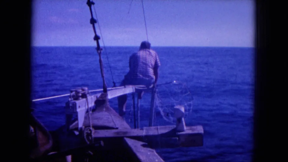 1964 Hawaii home-movie frame showing a man in a checkered shirt at a small boat bow with fishing net and rigging over blue sea.