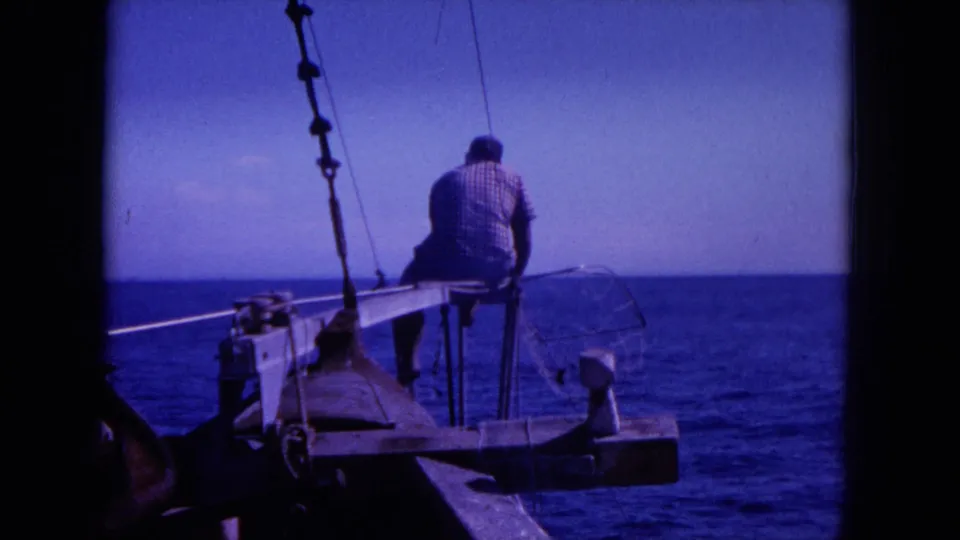 Home-movie frame from 1964 Hawaii showing a man seated at the boat edge with rigging and blue water in view.