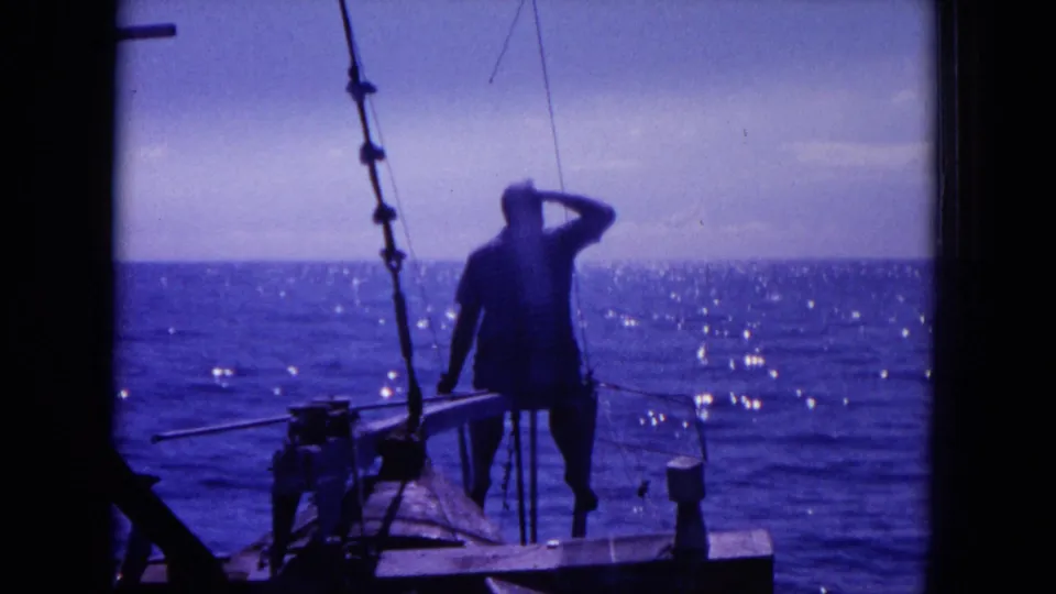 1964 Hawaii 8mm frame showing a man on a small fishing boat shading his eyes toward the open ocean.