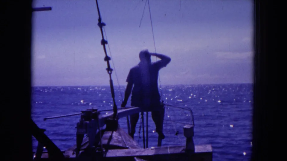 1964 Hawaii home-movie frame with a man beside fishing gear, shielding his eyes as the sea glints behind him.