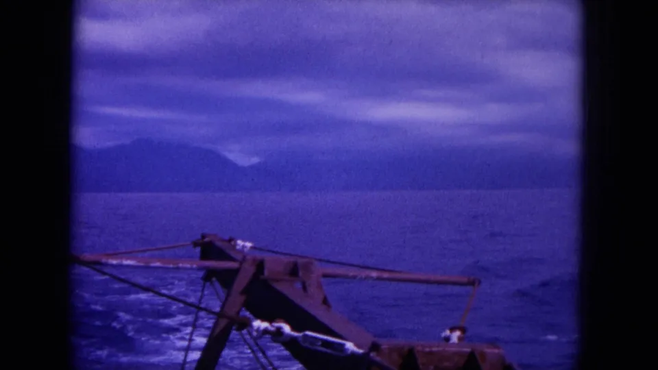 1964 8mm frame off Oahu with a deck-mounted harpoon gun in the foreground and ocean, wake, and mountains beyond.