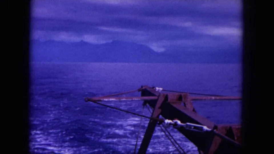 1964 Oahu home-movie frame showing a harpoon gun on a boat, rippled blue sea, wake, and a hazy mountainous shoreline.