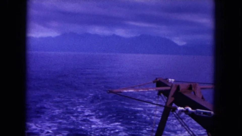 1964 8mm frame from off Oahu showing the harpoon gun at frame edge, open sea, boat wake, and distant coastal mountains.