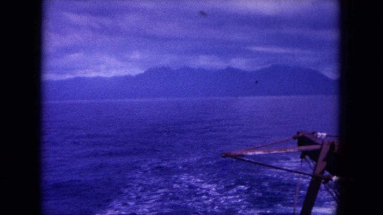 1964 8mm-style view likely off Hawaii, showing open water, boat wake, distant shoreline, cloudy sky, and part of the boat at right.